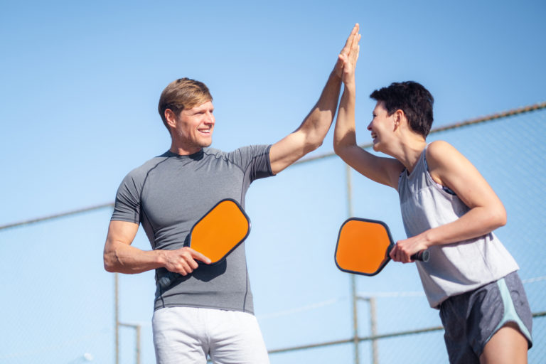 Laughing couple playing pickleball game, hitting pickleball yellow ball with paddle, outdoor sport leisure activity, celebrating victory 